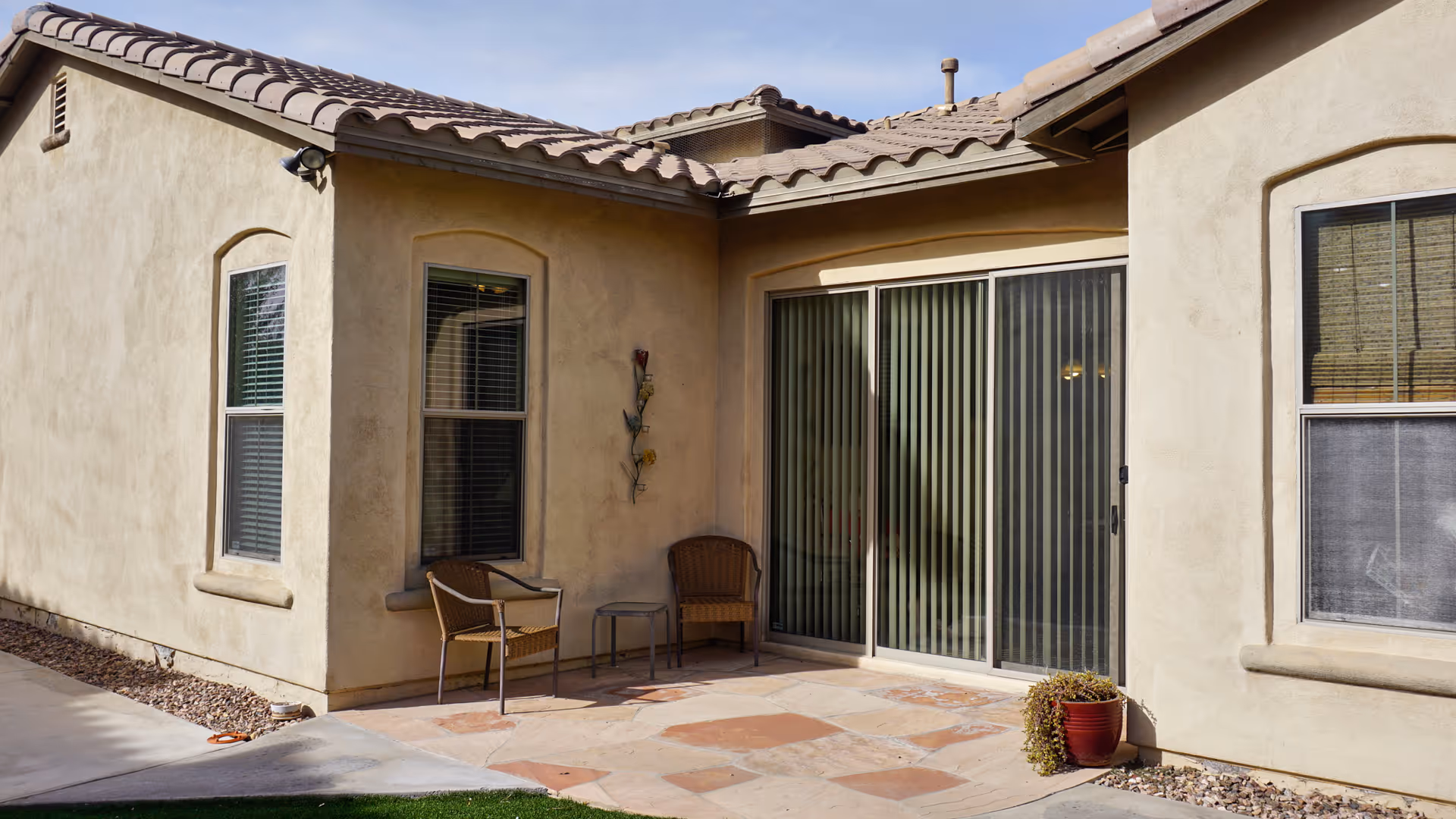 Outdoor patio area of a stucco senior living unit with a sliding glass door, two chairs, and a potted plant.