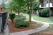 Curved concrete walkway through a landscaped courtyard with trees, shrubs, and an apartment-style building in the background.