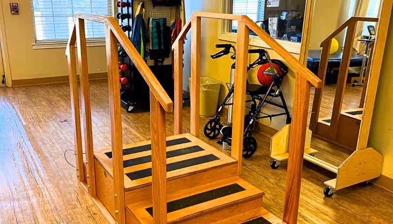 A small wooden exercise staircase with black anti-slip strips on the steps, located in a room with wooden flooring. Behind the staircase, there is a walker, a large mirror on wheels, and some exercise equipment including a rack with weights and resistance bands. The room has windows with blinds and yellow walls.