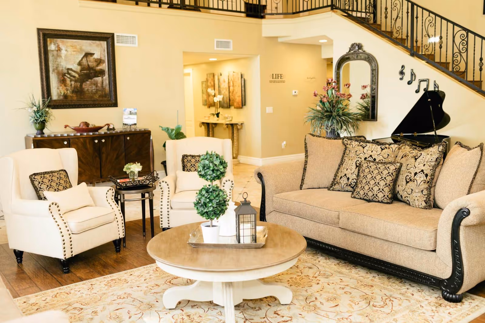 A cozy living room with a beige sofa adorned with patterned cushions, two cream armchairs with decorative pillows, a round wooden coffee table with plants and a lantern, a wooden sideboard with plants and decor, a framed painting of a piano on the wall, and a black grand piano under a staircase with musical note wall decorations.