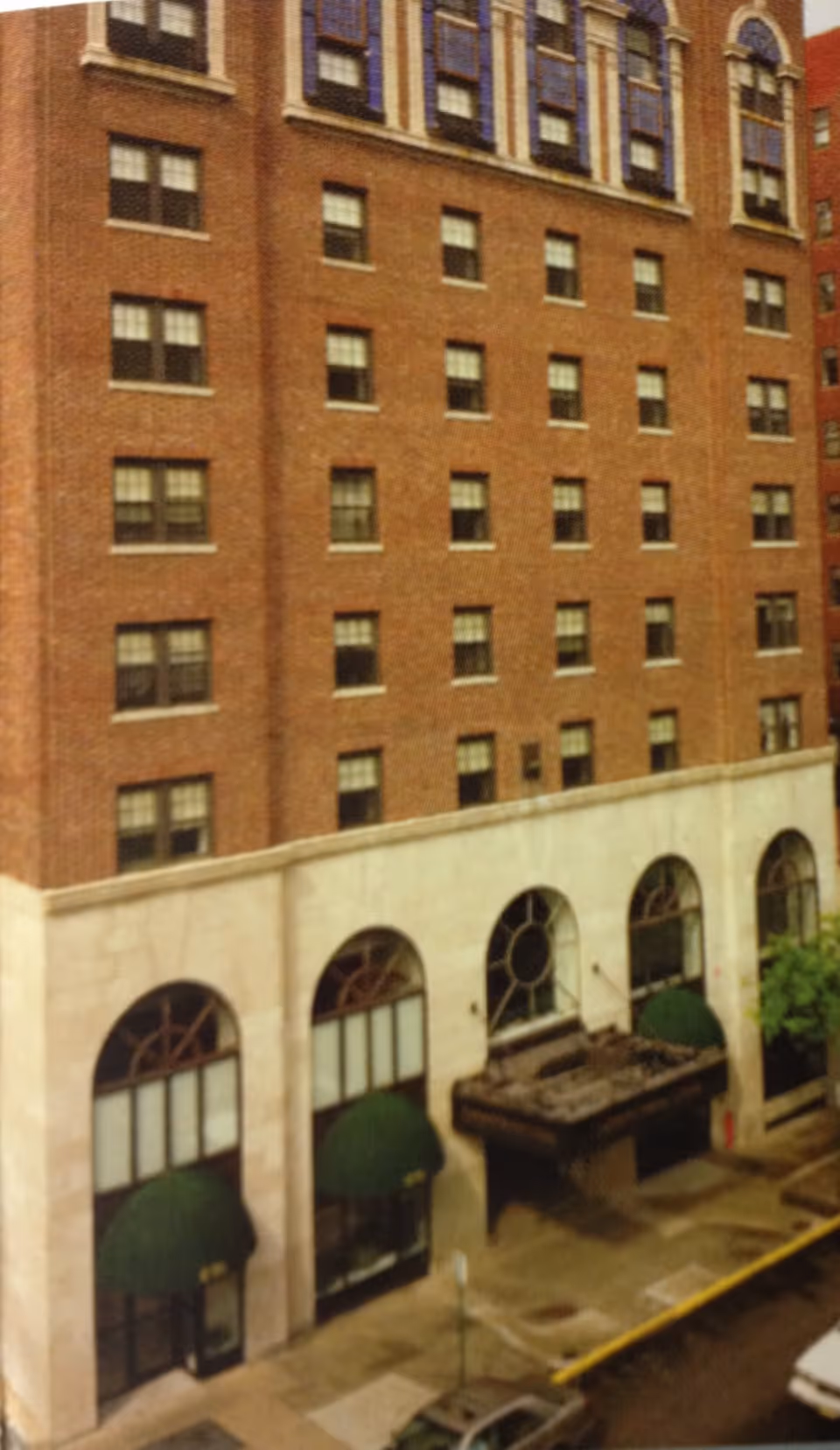 Exterior view of a multi-story brick building with large arched windows on the ground floor, green awnings above the entrances, and a small canopy over one entrance. The building is located on a city street with parked cars visible.