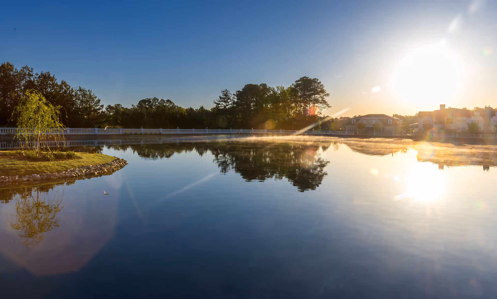 A serene outdoor scene at sunrise with a calm pond reflecting trees and buildings in the background, surrounded by a white fence and greenery.