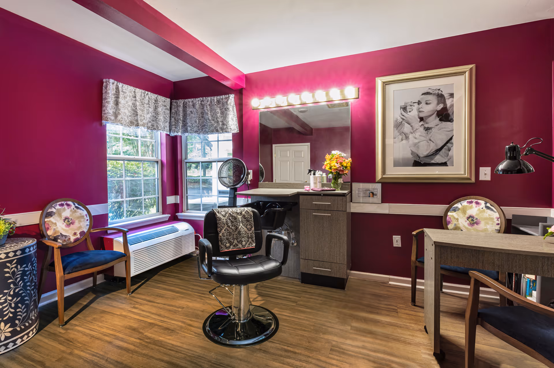 A bright salon room with deep magenta walls featuring a black salon chair in front of a large mirror with vanity lights. There are two floral-patterned chairs, a wooden desk with a black lamp, and a framed black-and-white photo of a woman applying makeup on the wall. The room has two windows with patterned valances and a wooden floor.