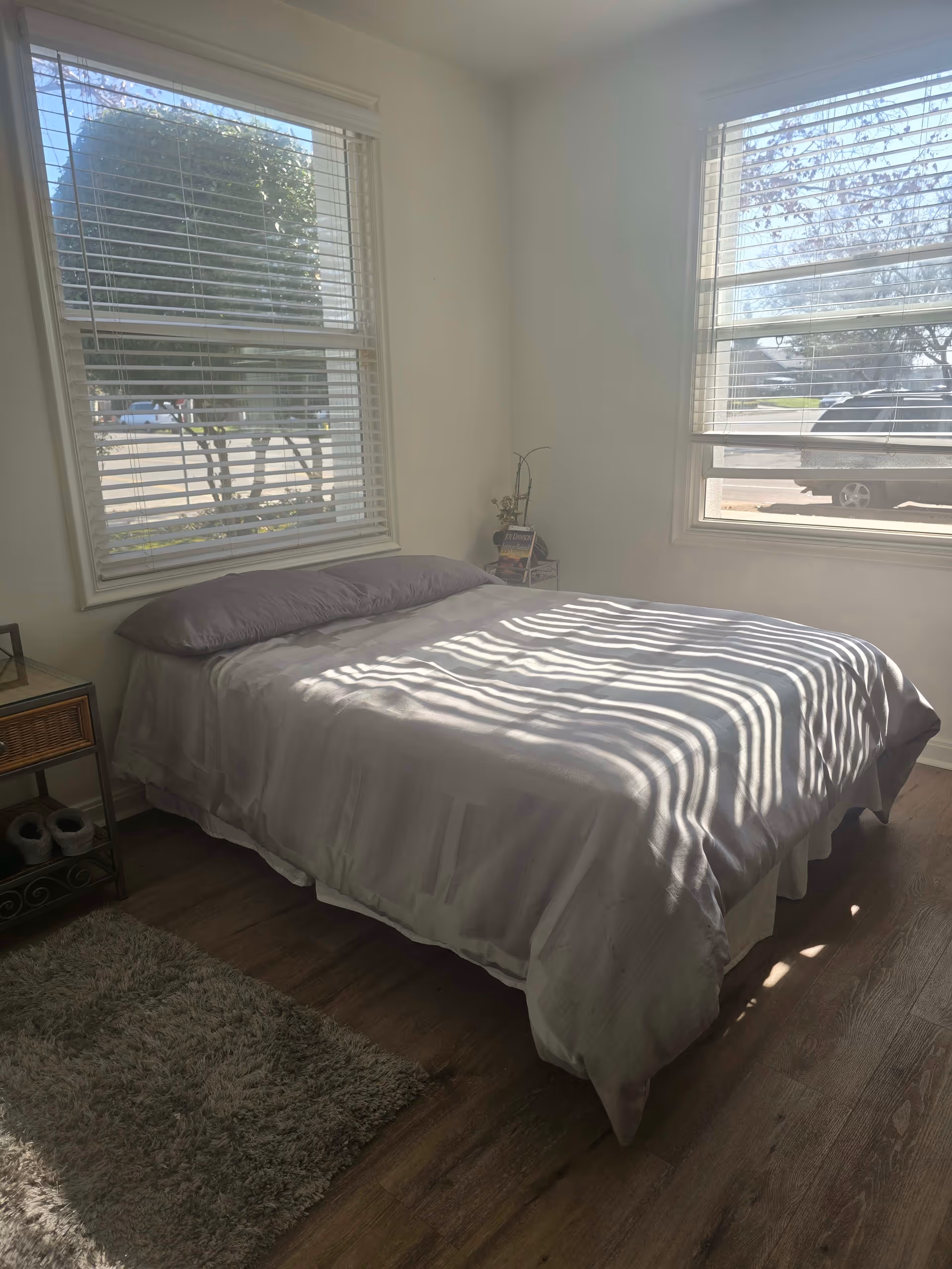 A bright bedroom with a neatly made bed covered in white bedding and two pillows. The room has two large windows with white blinds allowing sunlight to cast striped shadows on the bed. There is a small bedside table with a few books and a decorative plant, a small rug on the wooden floor, and slippers underneath a small metal and wood shelf.