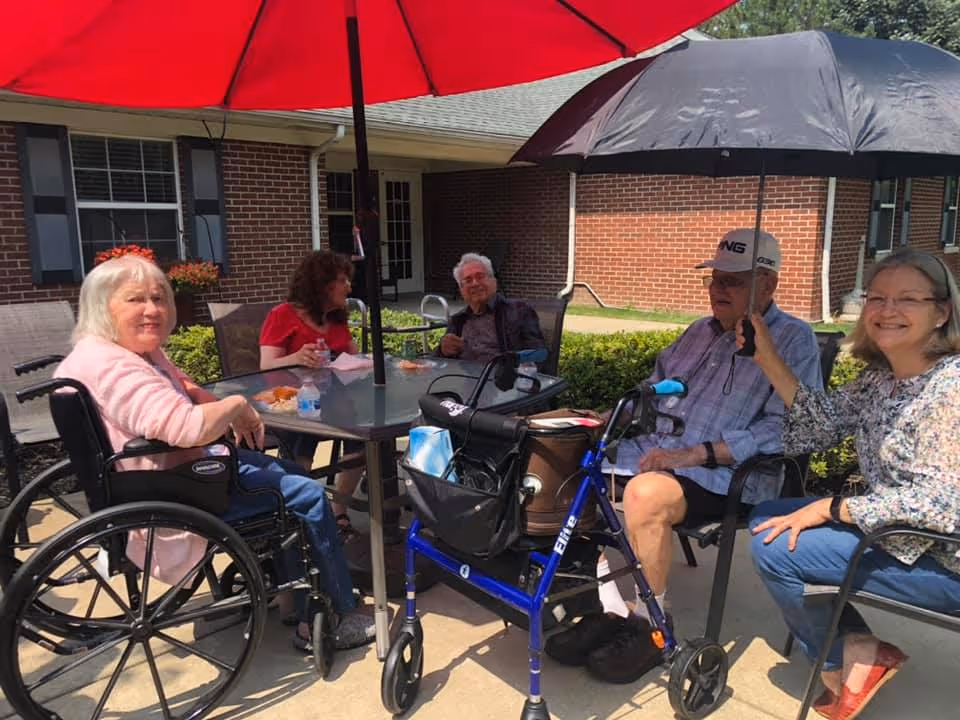 Five elderly people sitting outdoors around a glass table under a red umbrella. One woman in a wheelchair is smiling at the camera, another woman is holding a black umbrella over an elderly man wearing a cap. They are seated on a patio with brick buildings and greenery in the background.