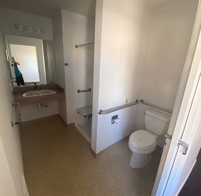 A clean, accessible bathroom with a toilet equipped with grab bars on both sides, a shower area with a fold-down seat and grab bars, and a sink with a mirror above it. The walls and floor are light-colored, and the space appears well-lit.