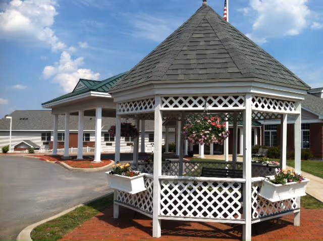 A white wooden gazebo with a gray shingled roof and hanging flower baskets, situated on a brick-paved area near the entrance of a building with a covered driveway and green roof. The sky is partly cloudy.