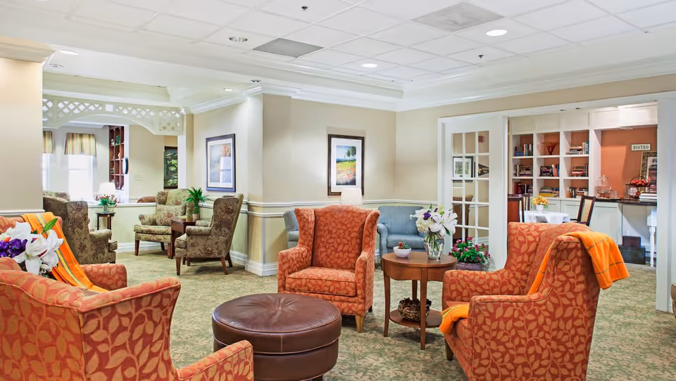 Bright, well-lit senior living lounge with upholstered armchairs arranged around a central ottoman and side tables, with bookshelves and a dining area in the background.