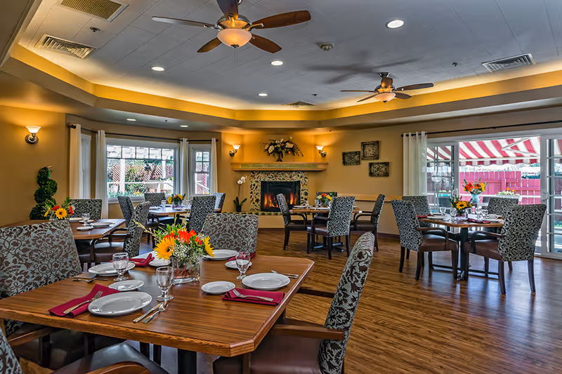 Spacious dining room with multiple set wooden tables, upholstered patterned chairs, floral centerpieces, ceiling fans, and a lit fireplace.