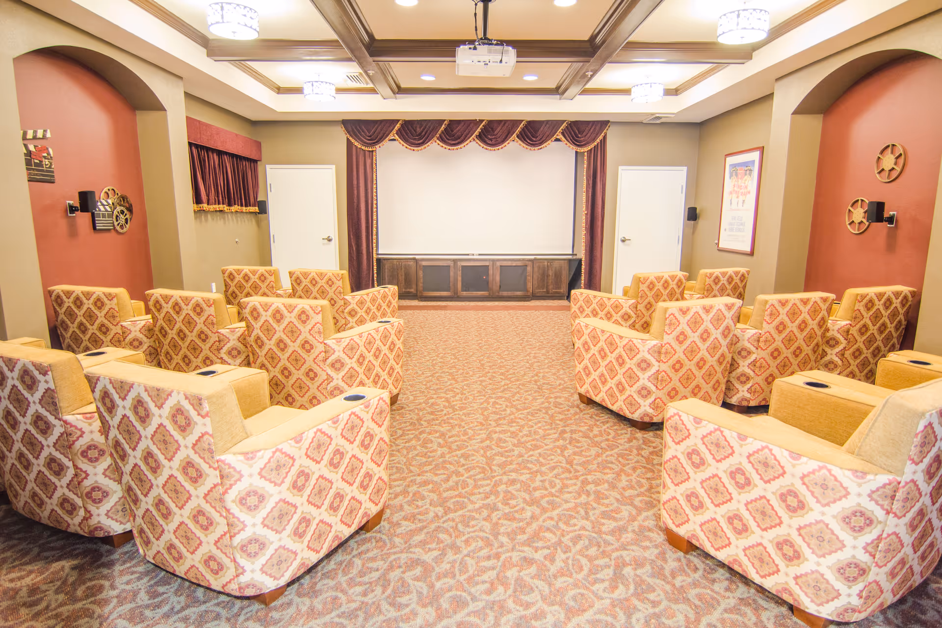A cozy theater room with patterned armchairs arranged in rows facing a large white projection screen framed by burgundy curtains. The room has beige walls with red accent panels, decorative film reels on the walls, and a patterned carpet. Ceiling lights and a projector are visible.