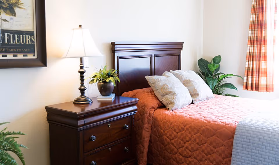 A cozy bedroom with a wooden headboard bed covered with an orange quilt and two decorative pillows. Next to the bed is a wooden nightstand with a table lamp and a small potted plant. A window with plaid curtains allows natural light into the room, and there is a framed picture on the wall.