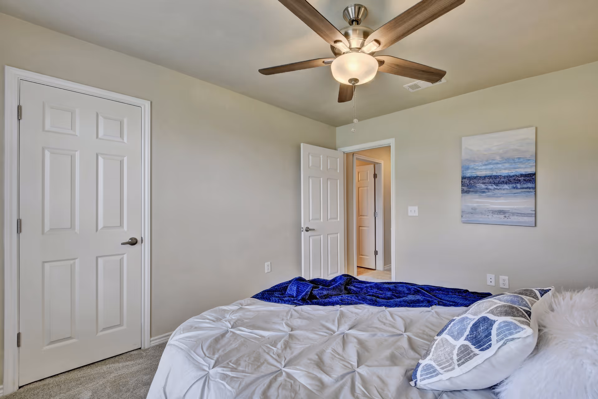 Bedroom with a bed dressed in white bedding and a blue throw, a ceiling fan, an open door, and coastal wall art.