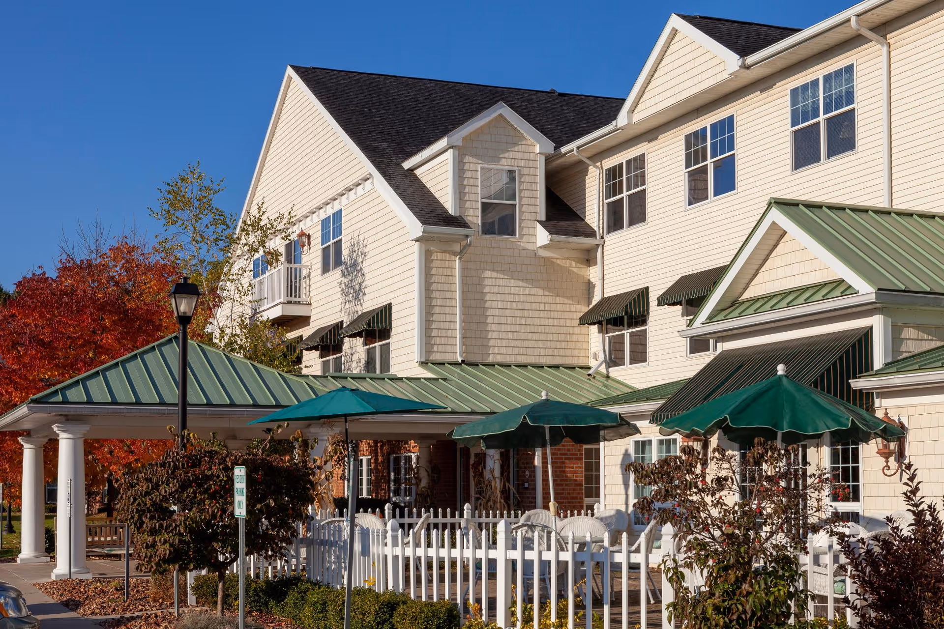 Exterior view of a senior living facility building with cream-colored siding and green metal roofs over entrances and patios. There are green umbrellas shading outdoor seating areas surrounded by white picket fences. Trees with autumn foliage are visible in the background under a clear blue sky.