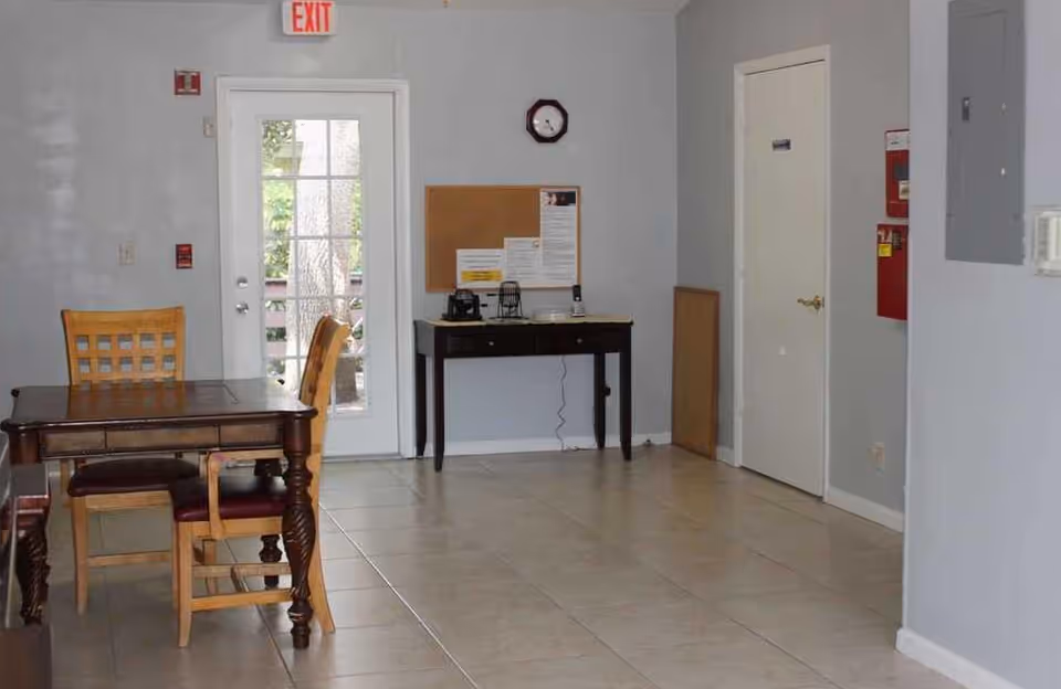 Interior room with light gray walls and tiled floor featuring a wooden table with three chairs on the left side. A glass door with an exit sign above it is centered on the back wall, next to a small black table with a corkboard and clock above it. A closed white door and a red fire alarm box are on the right wall.