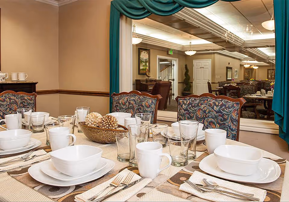 A dining table set with white dishes, glasses, and silverware in a decorated dining room with patterned chairs and a mirrored wall.