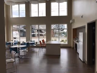 A spacious dining area with large windows letting in natural light. The room features several round tables with chairs arranged around them, and a kitchen area with white cabinets and appliances on the right side. The floor is wooden, and the walls are painted light beige.