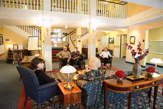 A bright and spacious common area in a retirement community with several elderly people sitting and conversing on comfortable chairs arranged around a central table with lamps and floral decorations. The room features a staircase with white railings, warm lighting, and a carpeted floor with a patterned rug.