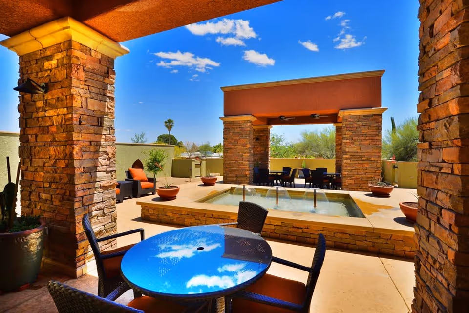 Outdoor patio area at Arroyo Gardens featuring a small rectangular water fountain surrounded by stone pillars and seating areas with tables and chairs under a clear blue sky with scattered clouds.