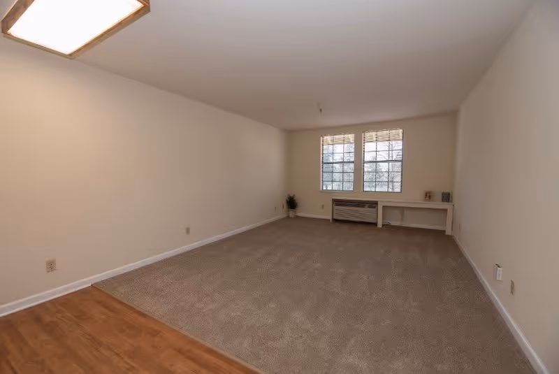 Empty carpeted living room with a small section of wood flooring, two windows with blinds and a low desk/bench beneath them.