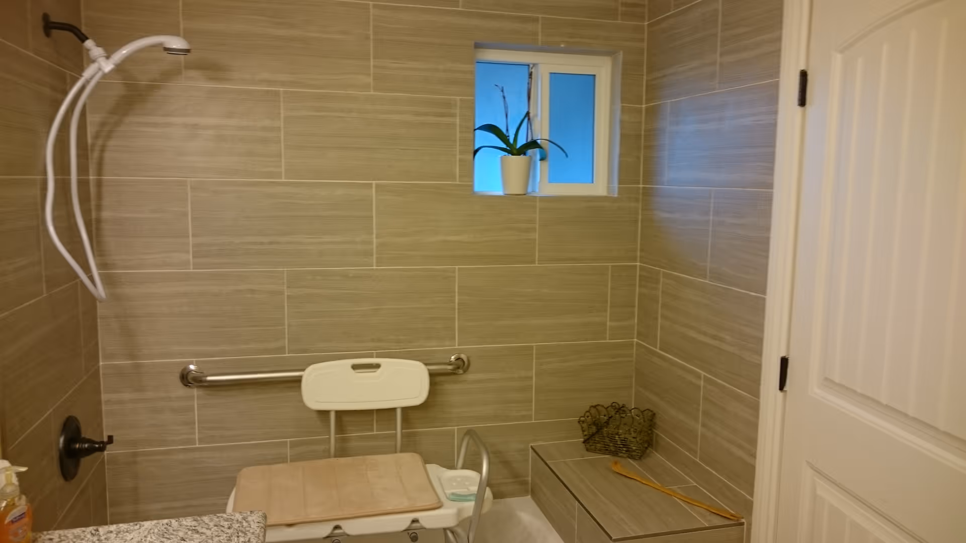 Bathroom with beige tiled walls featuring a white shower chair with a beige cushion, a handheld showerhead, a grab bar, a small window with a potted plant on the sill, a built-in bench with a basket and a wooden bath brush, and a white door.