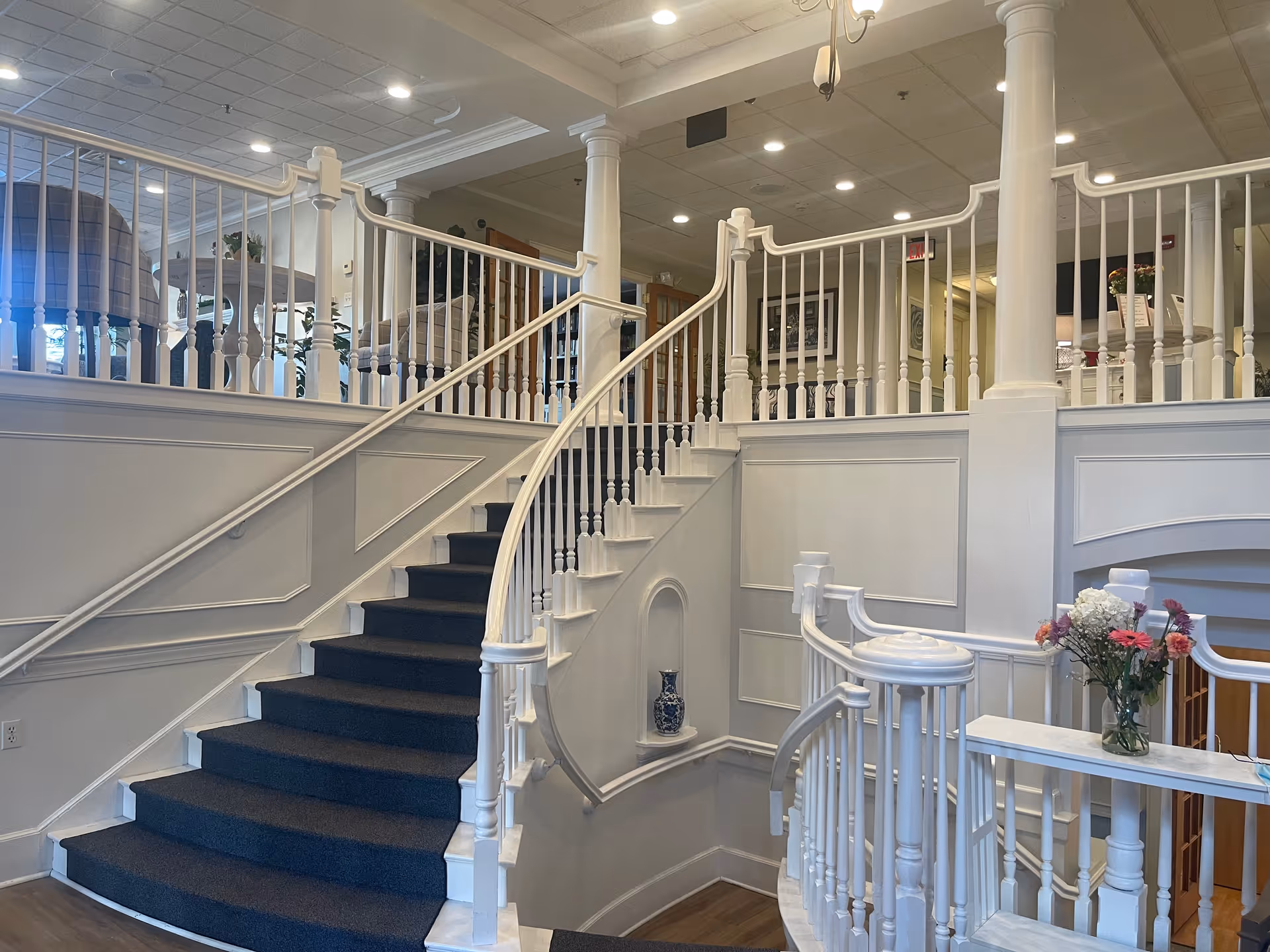 Elegant white interior lobby with a curved carpeted staircase, decorative railings, columns and a small vase of flowers on the banister.
