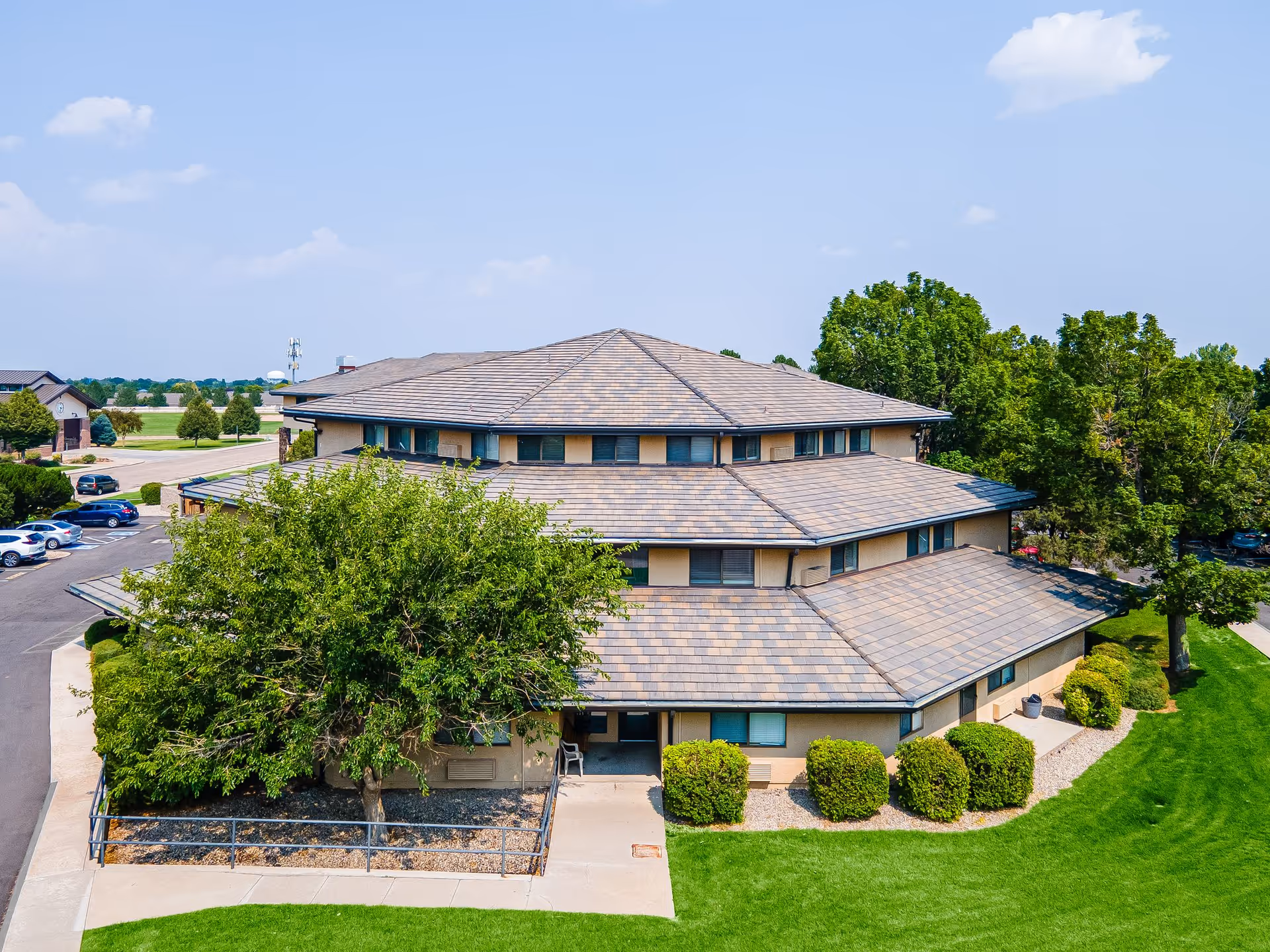 Exterior view of a multi-story building with a unique, tiered roof design surrounded by green trees, bushes, and a well-maintained lawn under a clear blue sky. There is a parking lot with several cars visible to the left.