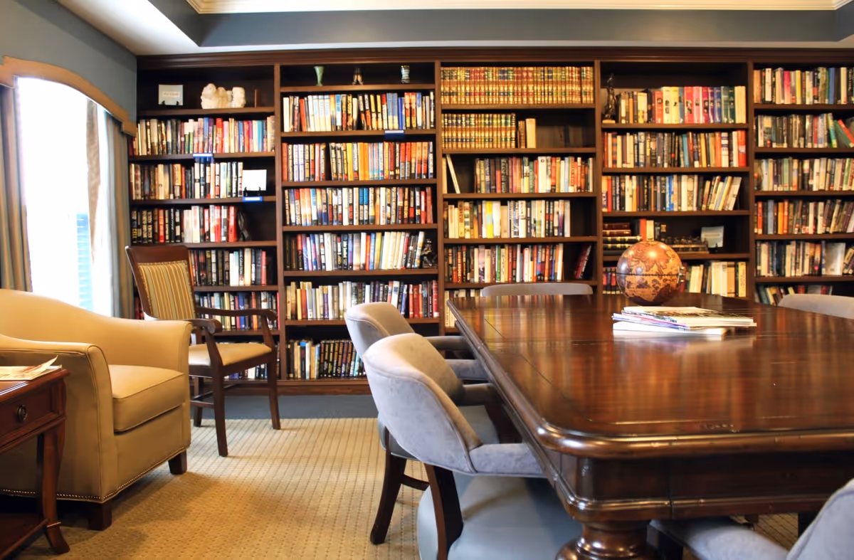 A cozy library room with wooden bookshelves filled with books along the back wall. In front of the shelves is a large wooden table surrounded by cushioned chairs. To the left, there is a beige armchair and a wooden side table near a window with curtains.