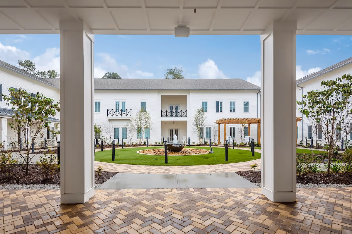 View from a covered patio area looking out onto a landscaped courtyard with a circular fountain in the center, surrounded by green grass, trees, and shrubs. The courtyard is enclosed by a two-story white building with balconies and windows, and a wooden pergola is visible on the right side.