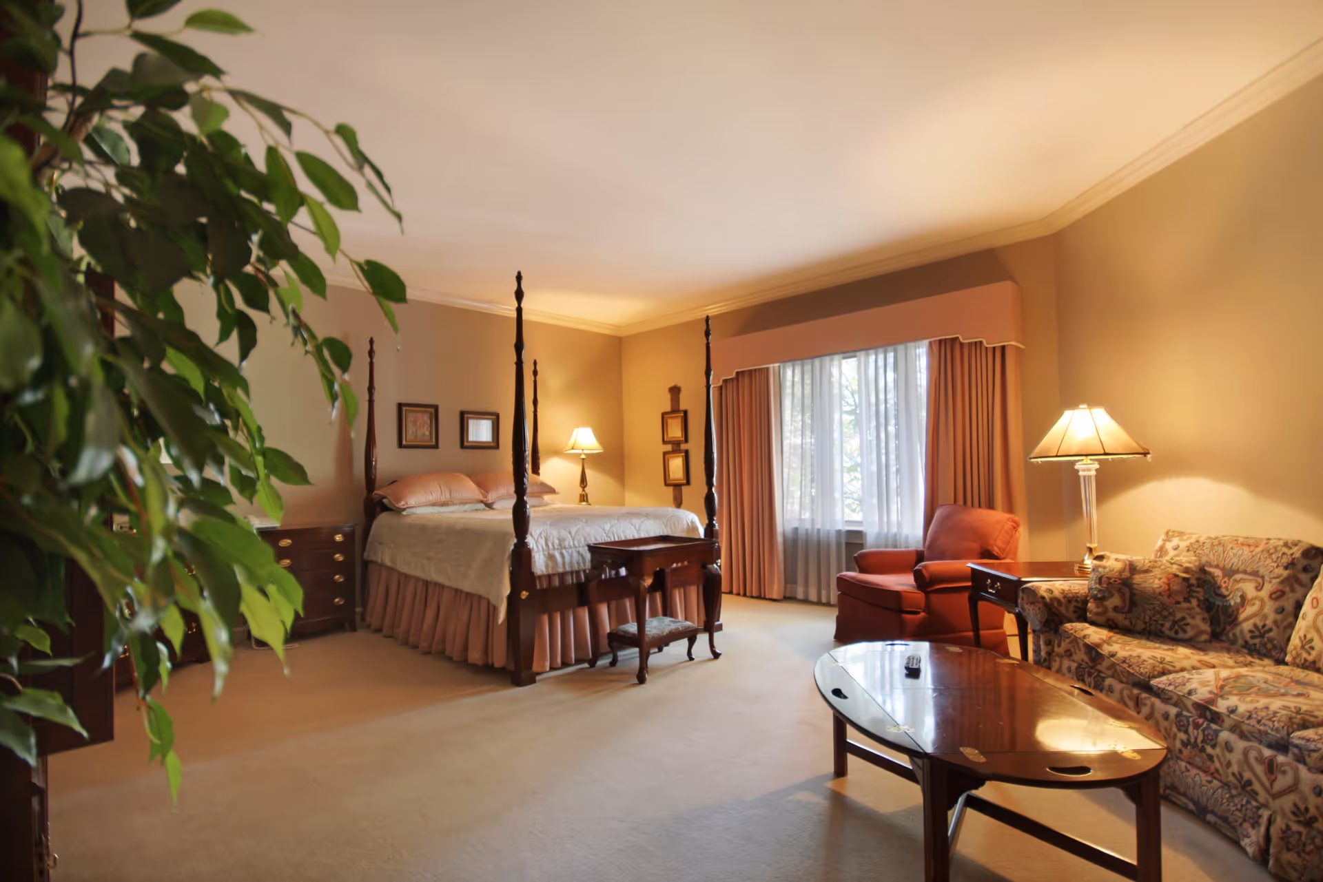 A spacious bedroom with a four-poster bed dressed in light bedding and pink pillows. There is a wooden nightstand with a lamp next to the bed. A large window with sheer white curtains and pink drapes lets in natural light. The room also features a patterned sofa, a red armchair, a wooden coffee table with a remote control on it, and a potted plant partially visible on the left side. The walls are painted beige, and the carpet is light-colored.