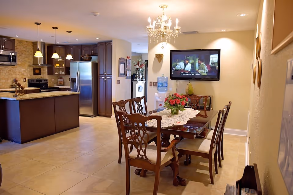 A dining area with a wooden dining table and six chairs, a floral centerpiece on a lace table runner, a wall-mounted TV showing a scene with two men, a water dispenser, and a chandelier overhead. Adjacent to the dining area is a kitchen with dark wood cabinets, stainless steel appliances including a refrigerator, microwave, and stove, and pendant lights hanging over a granite countertop island.