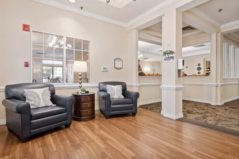 A cozy seating area in a senior living facility featuring two black leather armchairs with decorative pillows, a round wooden side table with a lamp and small plant, light beige walls, and a window looking into another room.