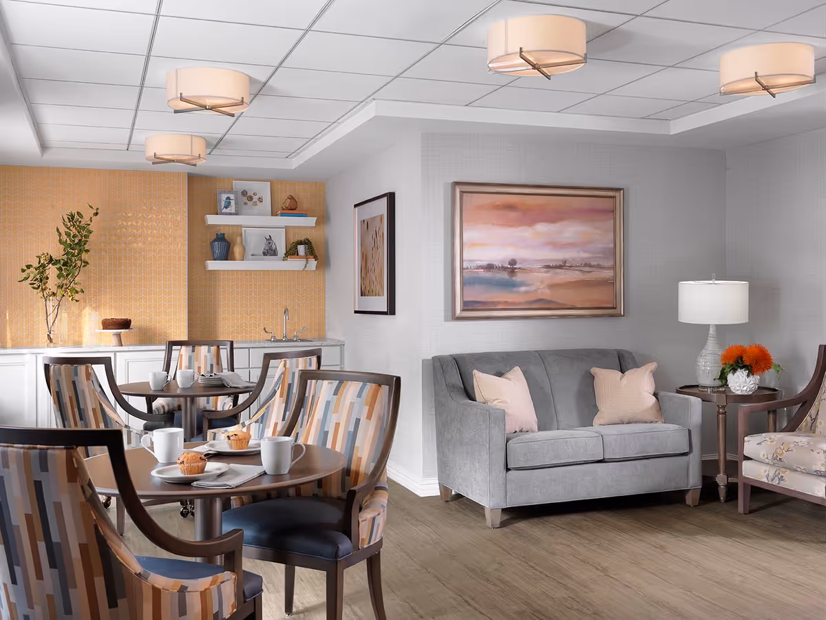A cozy common area in a senior living facility featuring a small round dining table with four patterned chairs, each set with white mugs and plates with muffins. In the background, there is a countertop with a small sink, decorative shelves with vases and framed pictures, and a plant. To the right, a gray loveseat with two light pink pillows sits next to a side table with a white lamp and a small floral arrangement. Above the loveseat hangs a framed landscape painting with soft pastel colors. The room has a warm, inviting atmosphere with soft lighting from ceiling fixtures.