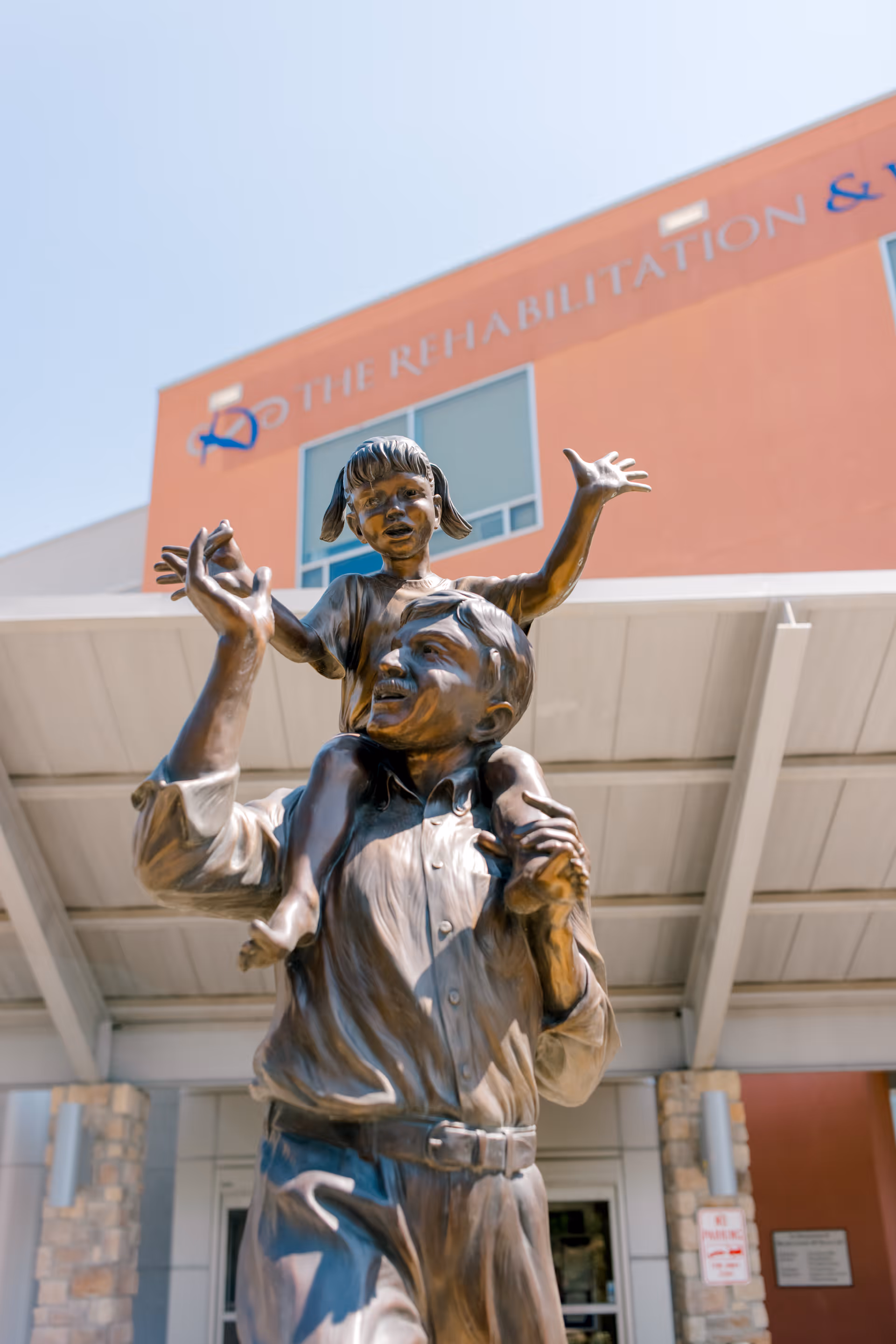 Bronze statue of a man carrying a young girl on his shoulders, both with joyful expressions, in front of The Rehabilitation & Wellness Centre of Dallas building.