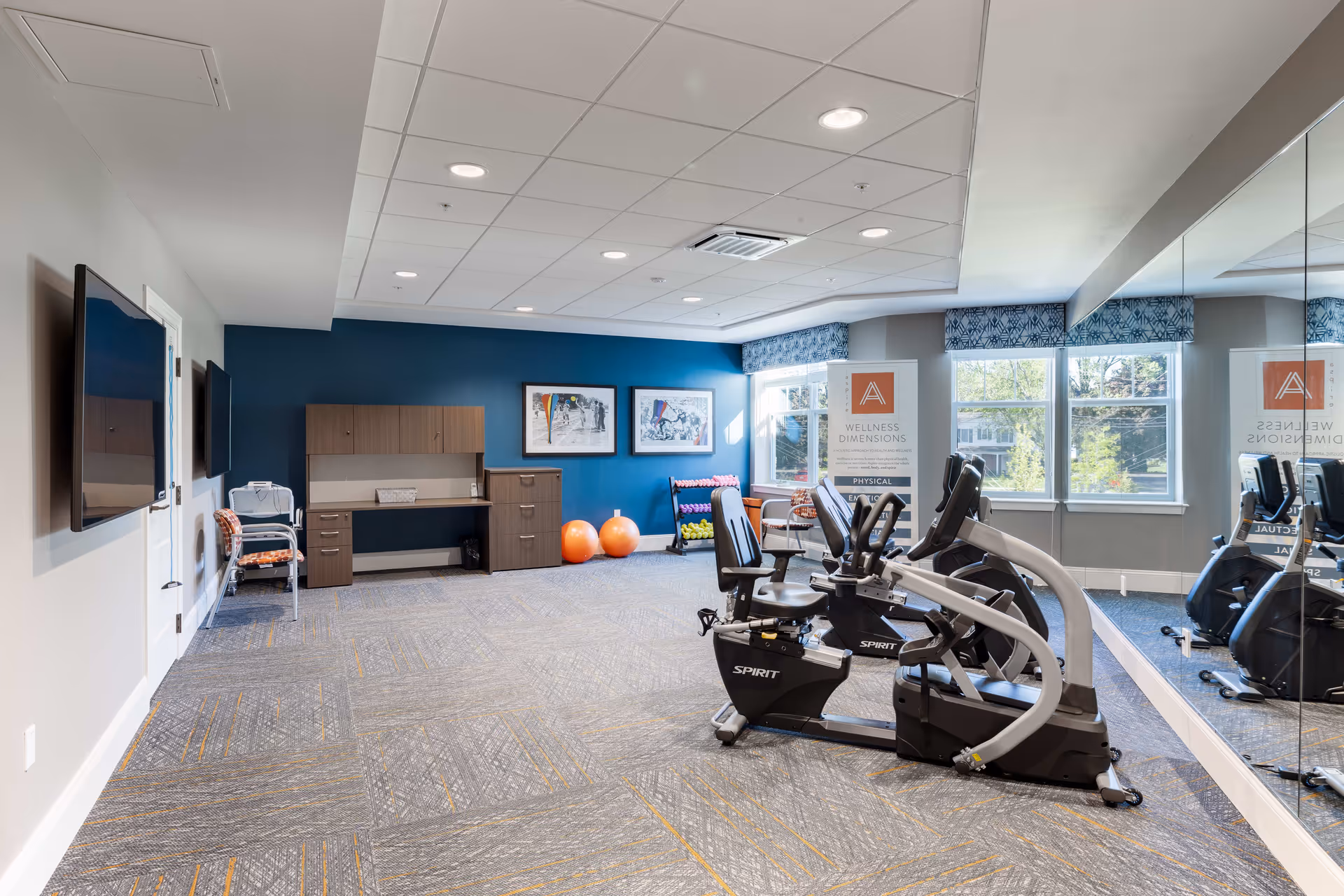 A bright fitness room with two recumbent exercise bikes, a large mirror on one wall, a blue accent wall with two framed pictures, a desk with cabinets, two orange exercise balls, a rack of colorful dumbbells, and a window with patterned valances letting in natural light.