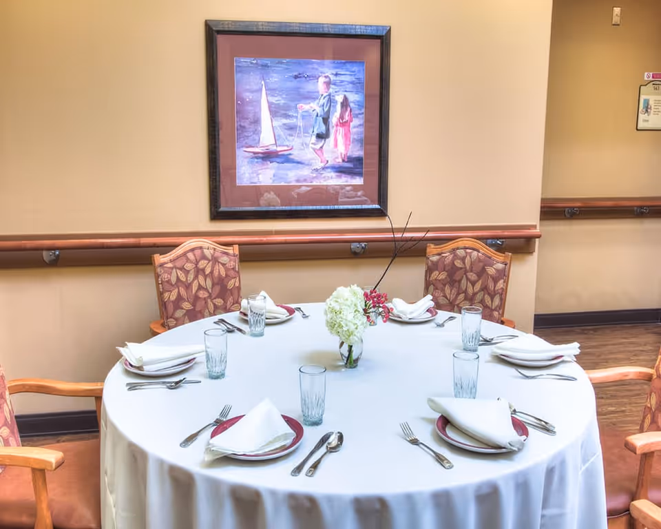 Round dining table set for six with white tablecloth, napkins, glassware and a floral centerpiece beneath a framed painting on a warm-colored wall.