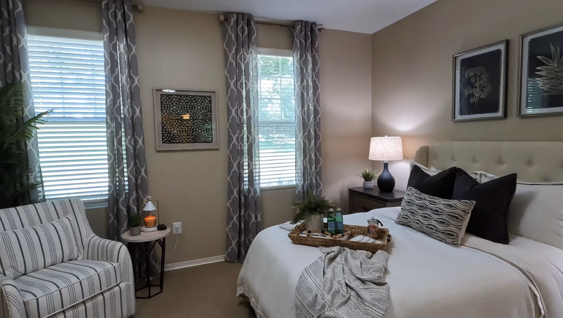 A cozy bedroom with a neatly made bed featuring white linens, black and patterned pillows, and a gray throw blanket. A wooden tray with water bottles and snacks sits on the bed. To the right of the bed is a nightstand with a lamp and a small plant. Two framed botanical prints hang on the beige wall above the bed. On the left side of the room, there is a striped armchair next to a small round table with a candle and a small plant. Two windows with patterned gray curtains allow natural light into the room.