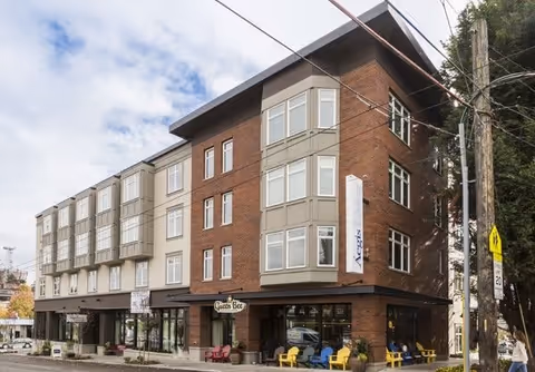 Exterior view of a multi-story senior living facility named Aegis Living Queen Anne Galer, featuring a combination of brick and light-colored siding with multiple windows. There are colorful chairs arranged outside near the entrance, and a vertical sign displaying the facility's name. The sky is partly cloudy.