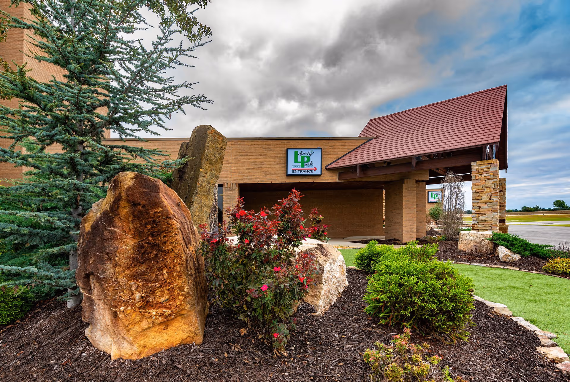 Exterior view of Liberty Park Assisted Living facility entrance with landscaped garden featuring large rocks, bushes, and a pine tree under a cloudy sky.