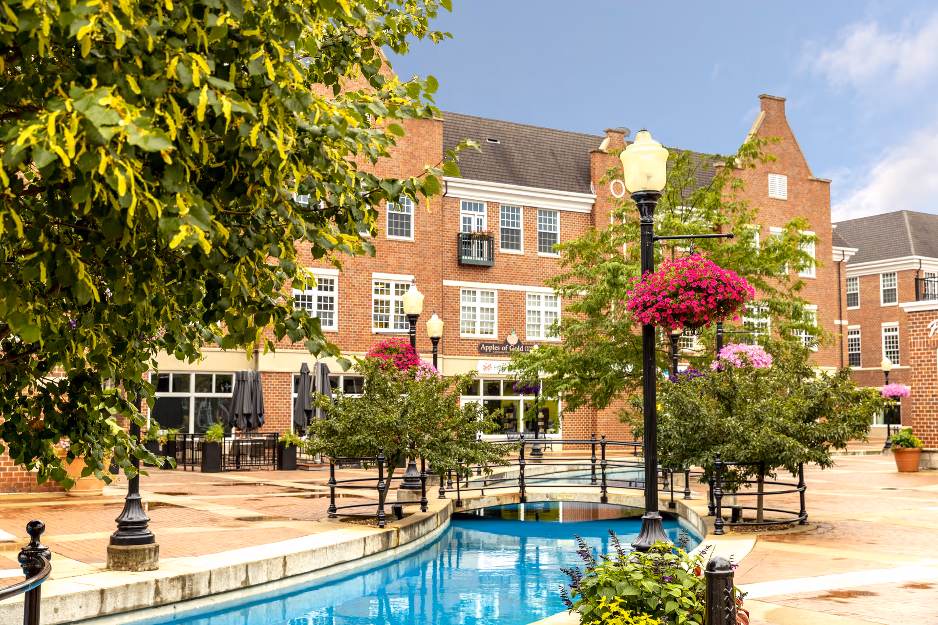 Outdoor courtyard area at Independence Village of Pella featuring a small water canal with a decorative bridge, surrounded by trees, hanging flower baskets on lamp posts, and a brick building with multiple windows in the background.