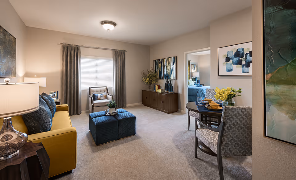 A cozy senior living room with a yellow sofa, blue cushions, a small armchair by a window with gray curtains, and two blue ottomans in the center. A wooden sideboard with decorative items and abstract paintings adorn the walls. A small round dining table with two patterned chairs and a vase of yellow flowers is visible near the entrance to a bedroom with a blue bedspread.