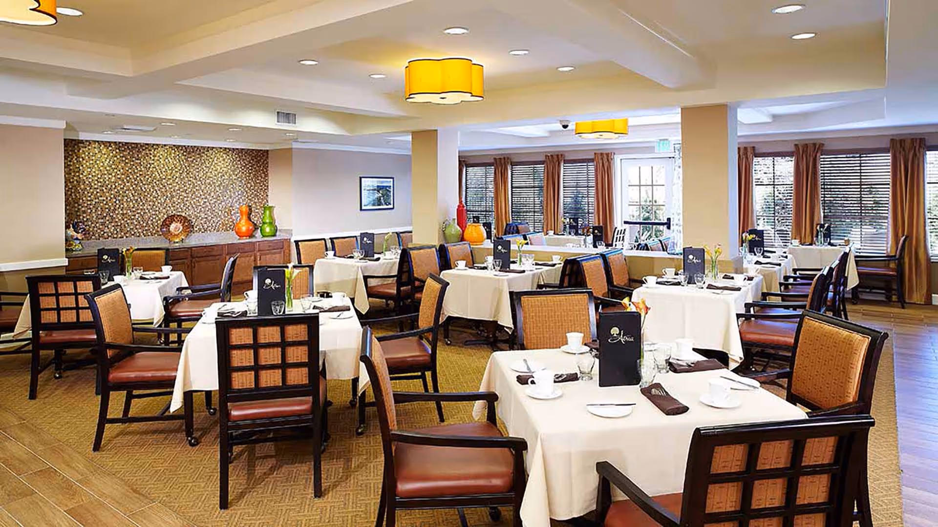 Dining room with neatly arranged tables covered in white tablecloths and wooden chairs under warm ceiling lights.