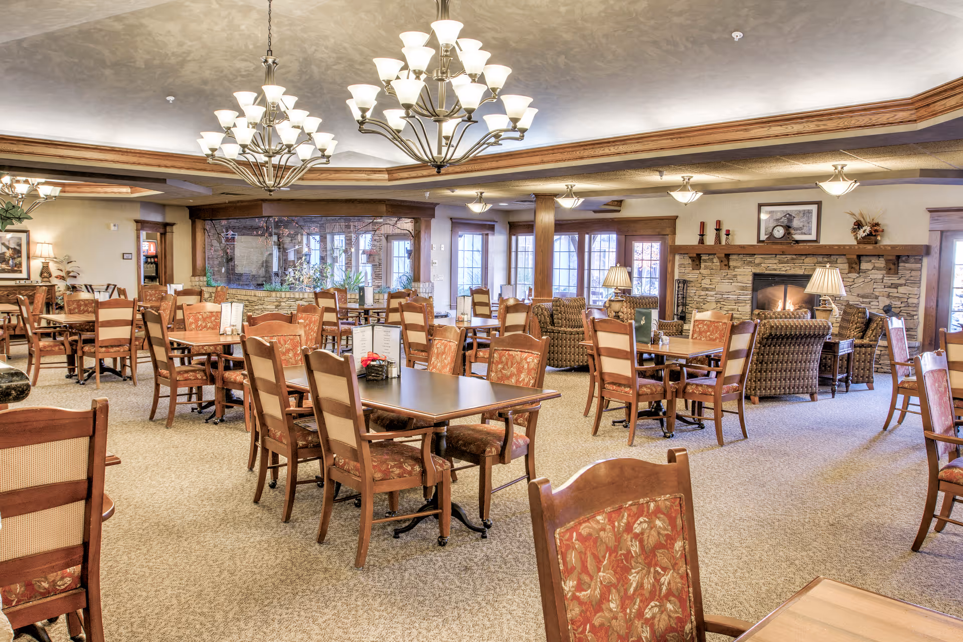 Spacious senior living dining room with multiple wooden tables and chairs, chandeliers, and a stone fireplace.
