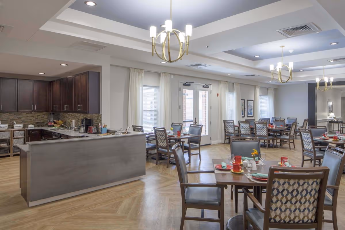A spacious dining room with multiple tables and chairs arranged neatly. Each table is set with red cups, plates, and napkins. The room has large windows with white curtains allowing natural light to enter. There are modern chandeliers hanging from the ceiling and a kitchen area with dark wooden cabinets and a countertop on the left side.