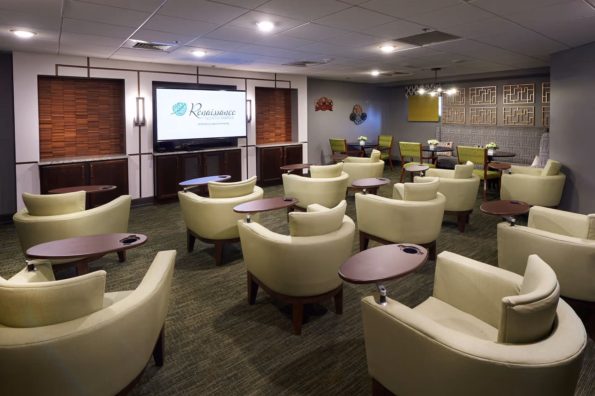 A cozy lounge area with multiple light green armchairs arranged in rows facing a large flat-screen TV mounted on the wall. Each chair has a small attached wooden table. The room has a green carpet, modern wall decor, and additional seating with green chairs and small tables in the back corner.