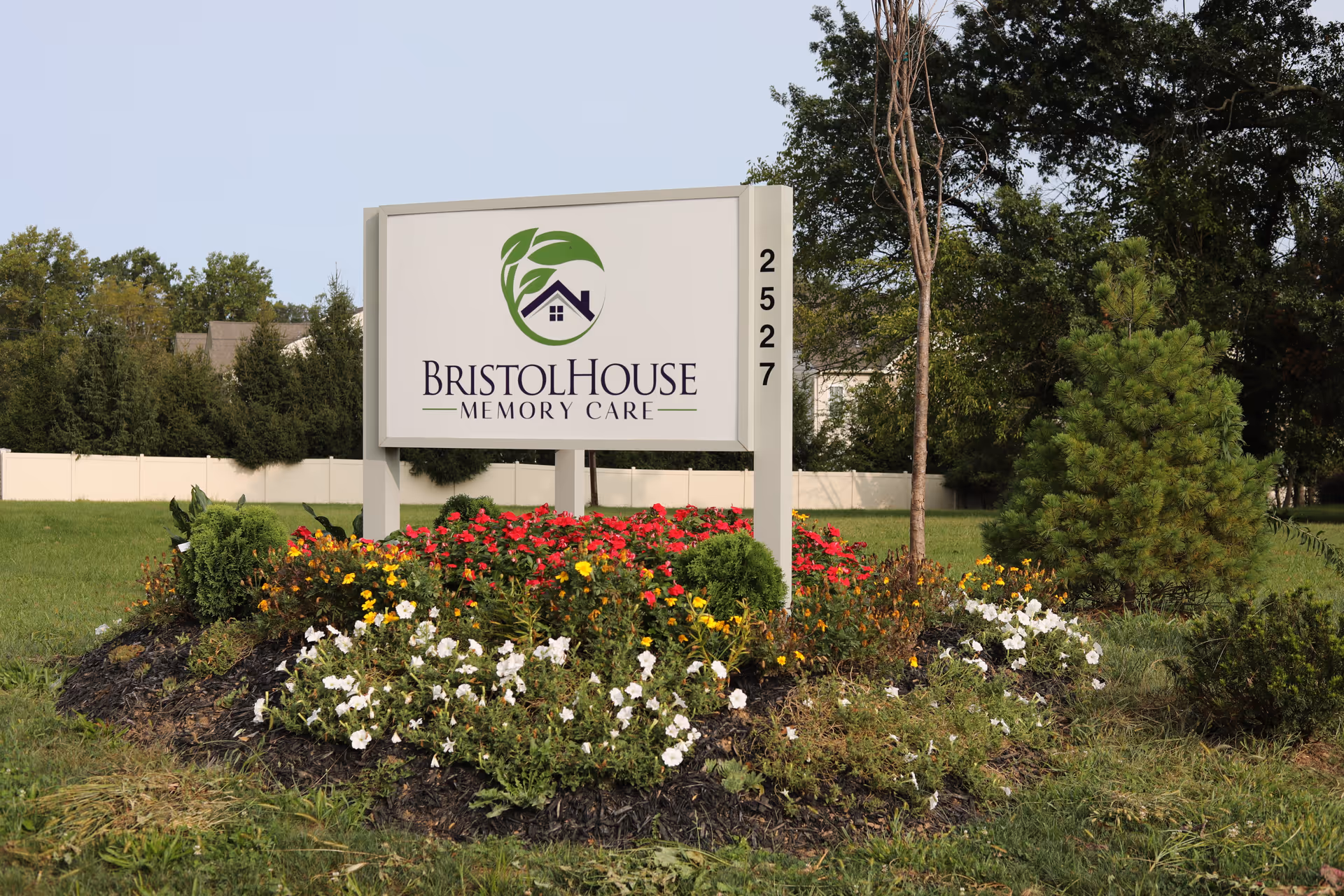 Outdoor view of a sign for Bristol House Memory Care surrounded by a landscaped flower bed with red, yellow, and white flowers, green shrubs, and trees in the background.