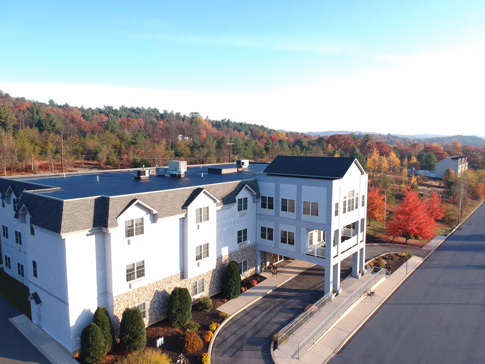 Aerial view of the front of a three-story senior living building with a covered entrance, driveway, landscaping and autumn trees.