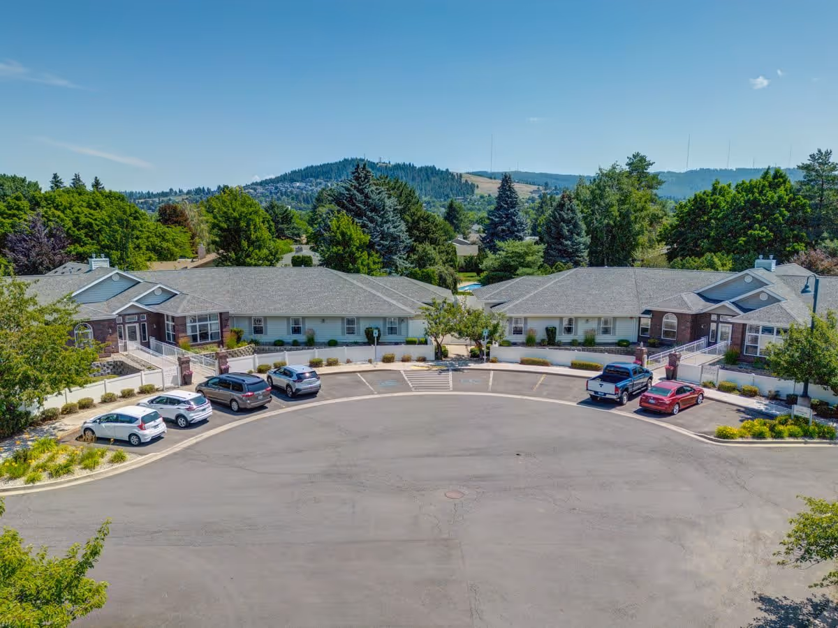 Aerial view of South Hill Village senior living facility showing two single-story buildings with gray roofs and brick accents, surrounded by trees and greenery. Several cars are parked in a semicircular driveway in front of the buildings, with hills and a clear blue sky in the background.