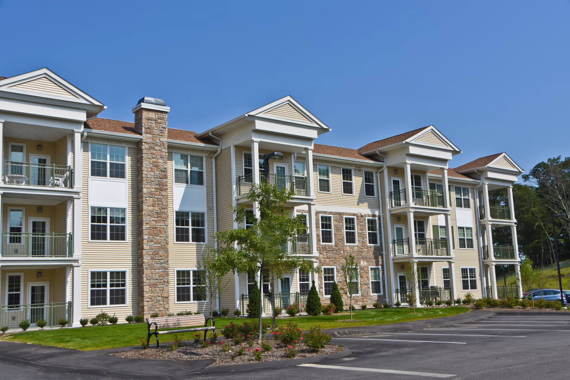 Three-story beige senior living building with balconies, stone accents, landscaped front and an empty parking lot under a clear blue sky.