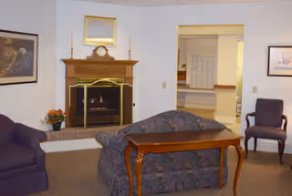 A cozy living room area featuring a lit fireplace with a wooden mantel, a floral-patterned couch facing away from the camera, a wooden table behind the couch, a purple armchair to the left, and another chair to the right. The walls are decorated with framed artwork, and there is a doorway leading to another room in the background.