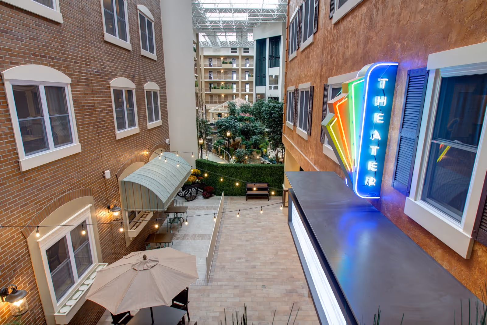 Indoor courtyard area of a senior living facility with brick and stucco walls, windows, string lights, outdoor seating with umbrellas, greenery, and a colorful neon sign that reads 'THEATER'.